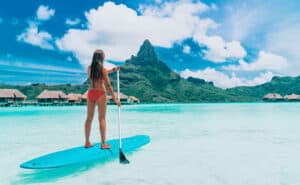 Woman standing up on paddle board, paddling over turquoise ocean at luxury beach resort hotel on Bora Bora island, French Polynesia