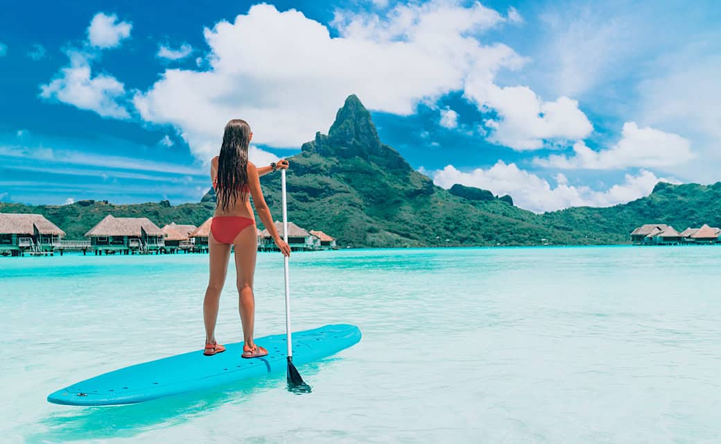 Woman standing up on paddle board, paddling over turquoise ocean at luxury beach resort hotel on Bora Bora island, French Polynesia