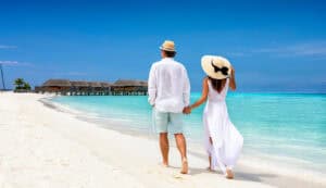 Happy couple in white clothing and with hats walks down a tropical beach with turquoise sea in the Maldives islands